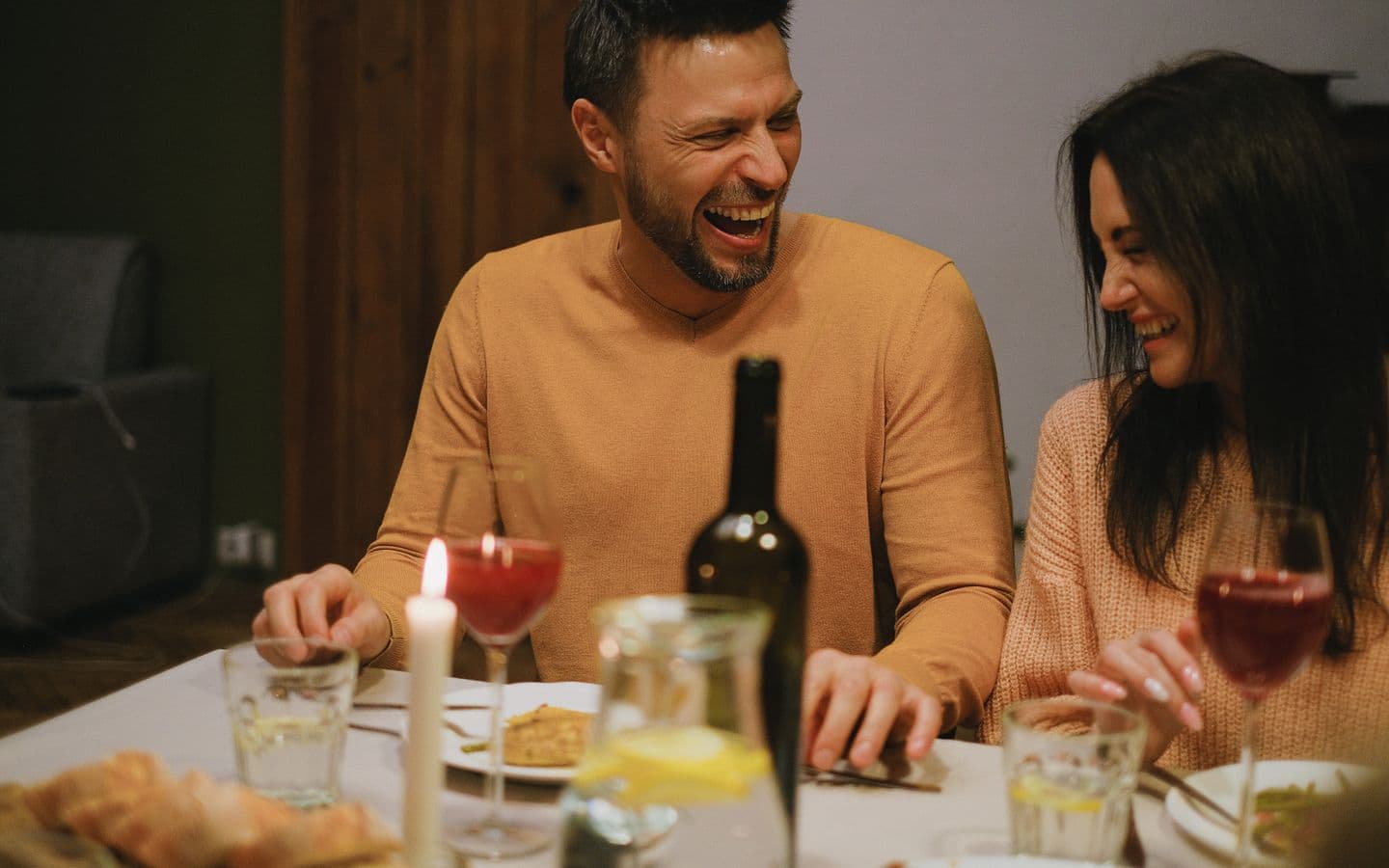 Fröhliches Paar lacht und schmust bei einem gemütlichen Abendessen mit Wein und Kerzenlicht