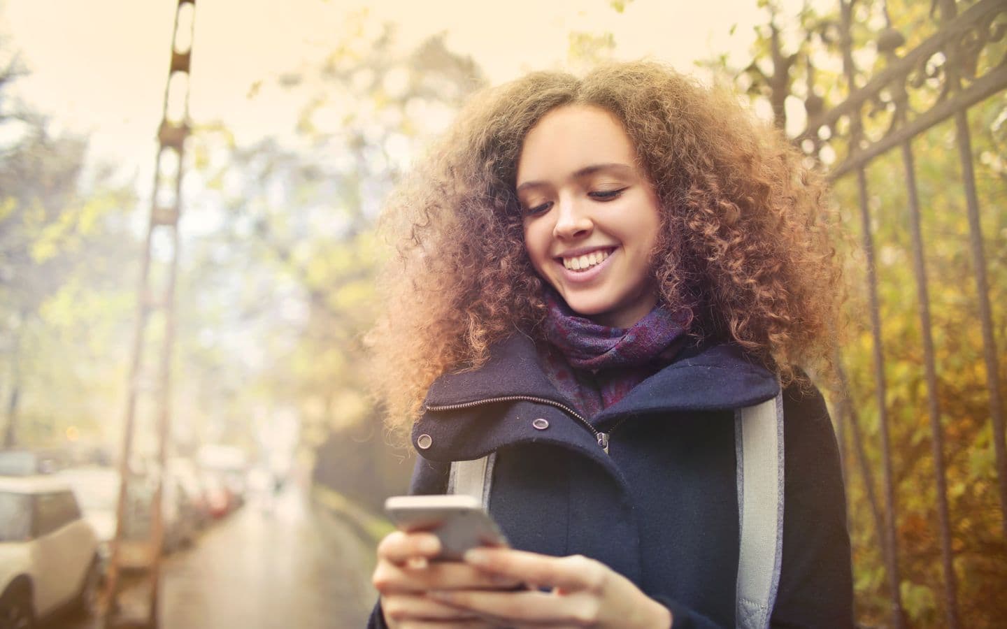 Junge Frau mit Locken lächelt beim Tippen auf ihrem Smartphone im herbstlichen Freien