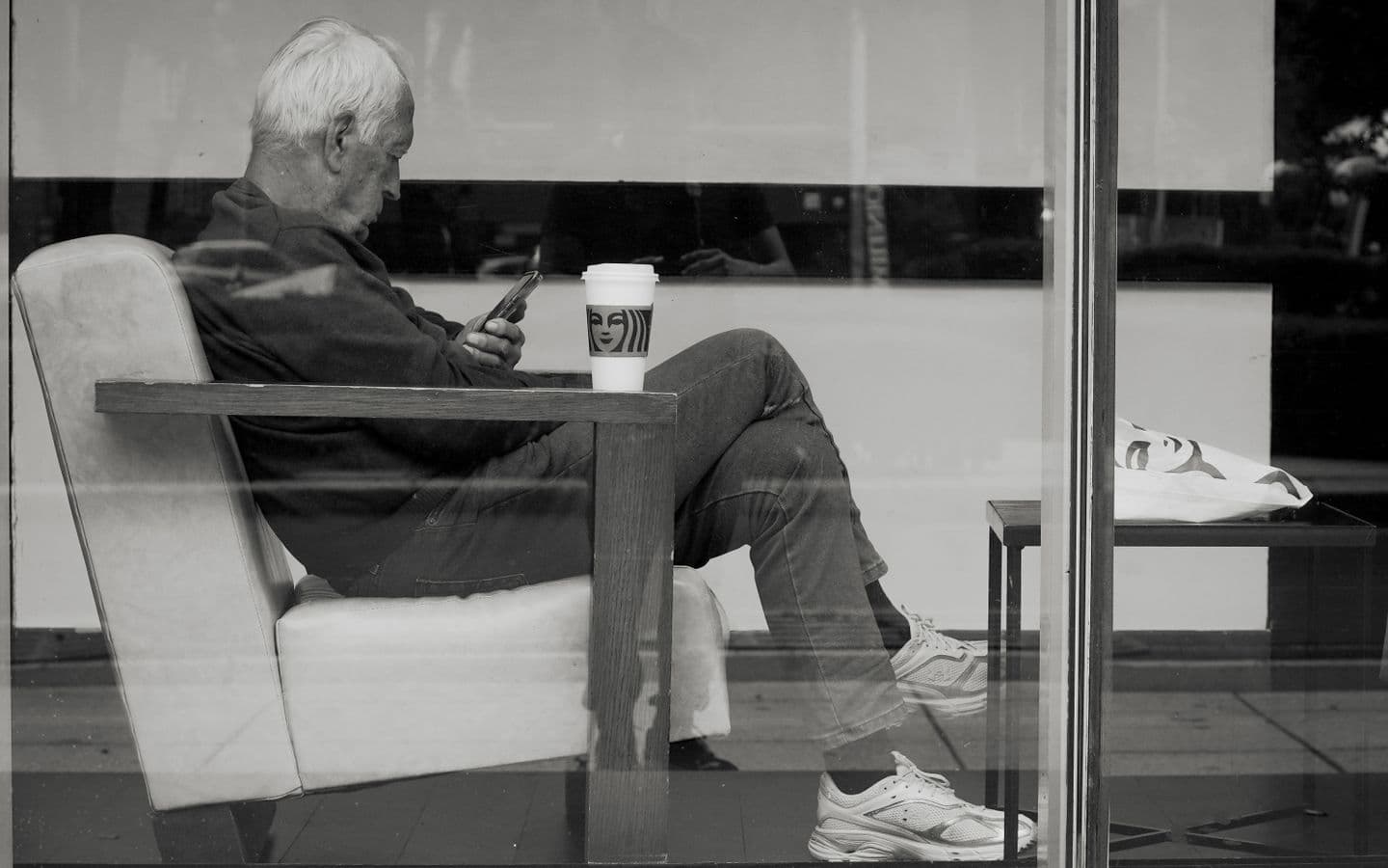 Älterer Mann sitzt mit einem Getränk in einem Café und schaut auf sein Handy