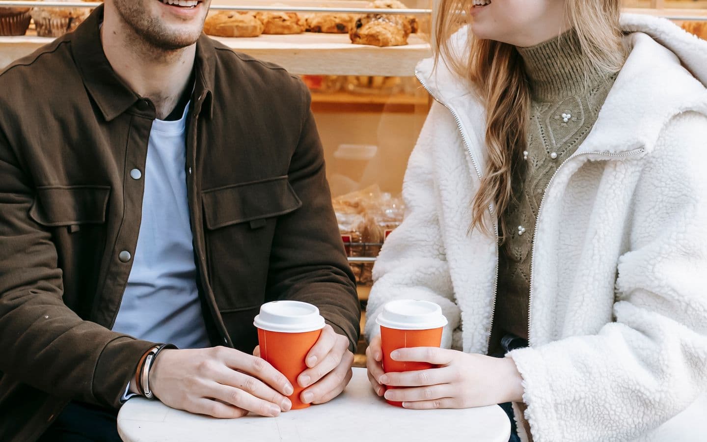 Glückliches Paar in Freizeitkleidung sitzt mit Heißgetränken in Pappbechern an einem kleinen Cafétisch