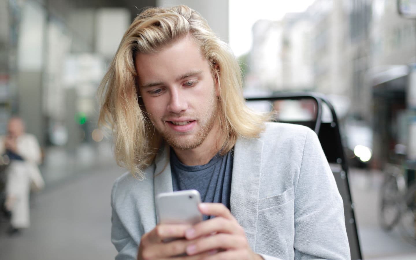 Lächelnder junger Mann mit langen blonden Haaren nutzt draußen in der Stadt sein Smartphone